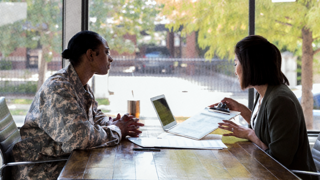 Two women sitting at a table near a window with a laptop between them. The black woman on the left is wearing an army uniform and the woman on the right is Asian and is holding a clipboard.