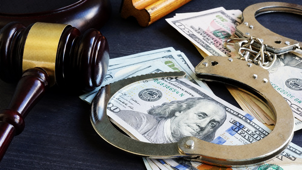 A close up of handcuffs laying on top of a one hundred dollar bill with a judge's gavel nearby.