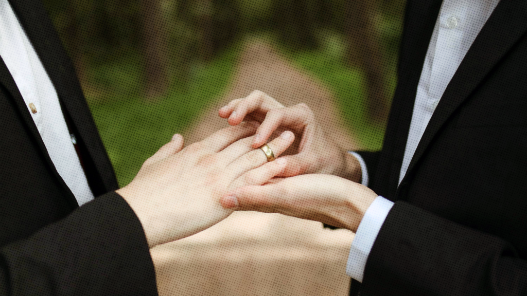 A close up of two white men's hands as they exchange wedding rings outdoors in a park.