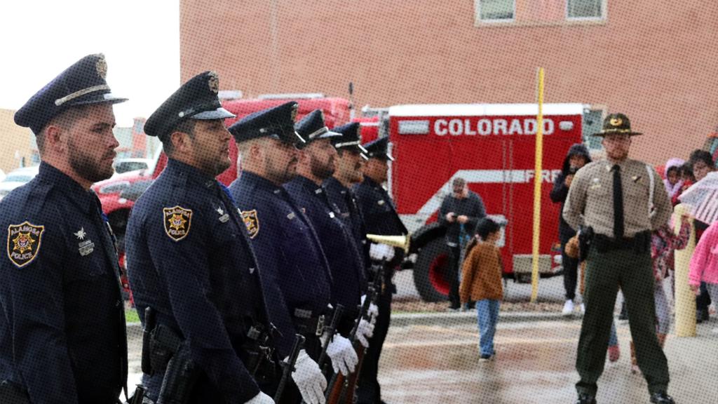 Six Alamosa police officers standing in line outside near a red firetruck with a sergeant facing the camera.