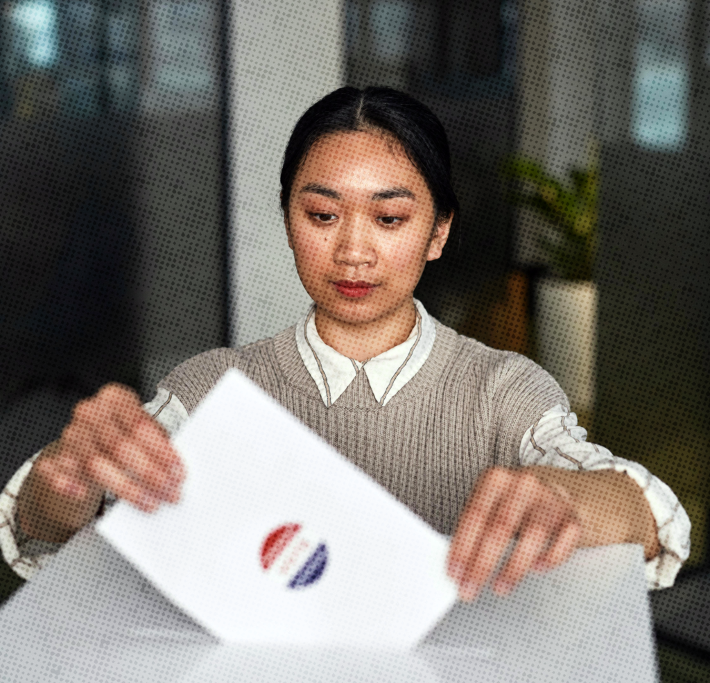 An Asian woman in an office with black hair and a collared shirt submitting a ballot into an election ballot box.