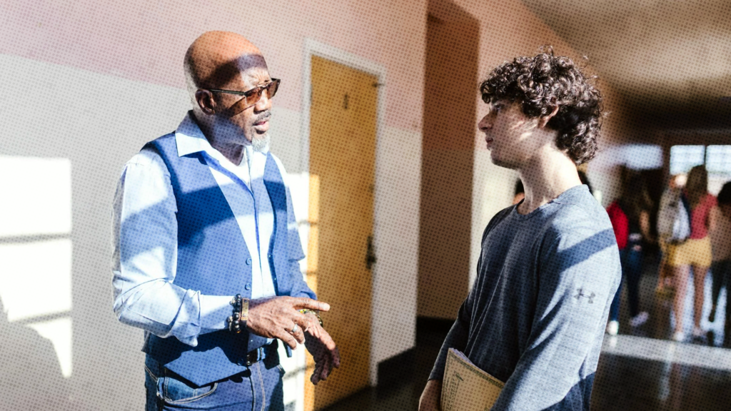 A black male teacher talking to a white male high school student in a school hallway.